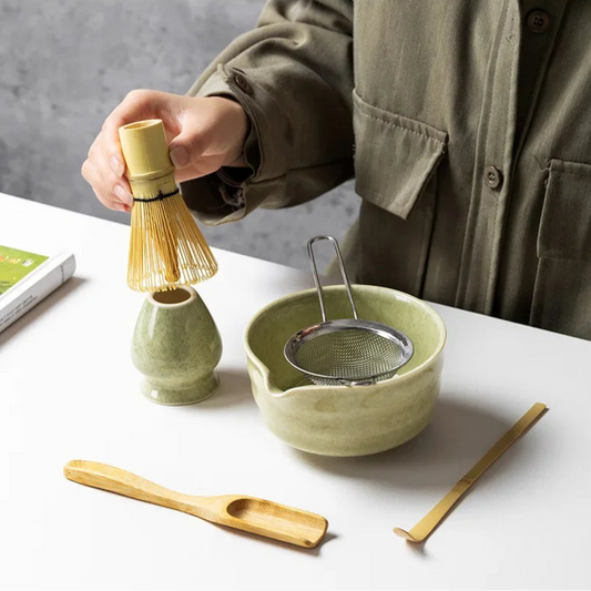 Person preparing tea with green ceramic bowl, whisk, and other tools on a white surface.