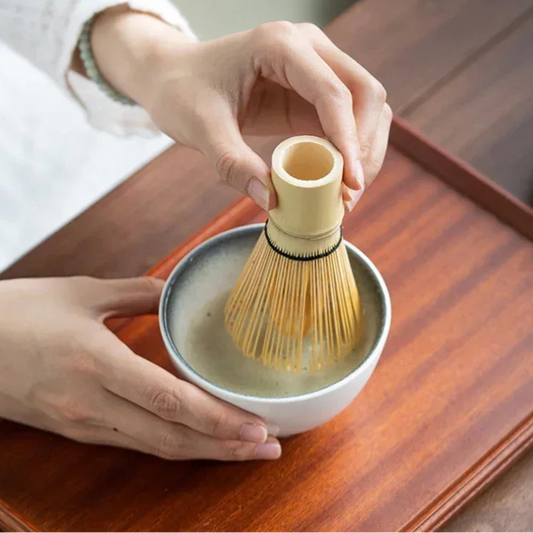 Person using a bamboo whisk to mix tea in a white bowl on a wooden tray, with 'Wontive' brand visible.