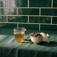 Brewed Tieguanyin oolong in a glass, pale gold, beside a gaiwan of unfurled leaves